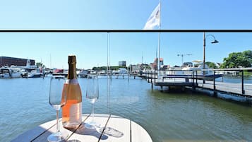 A bottle of champagne and two glasses on a wooden table by a body of water with boats and buildings in the background.