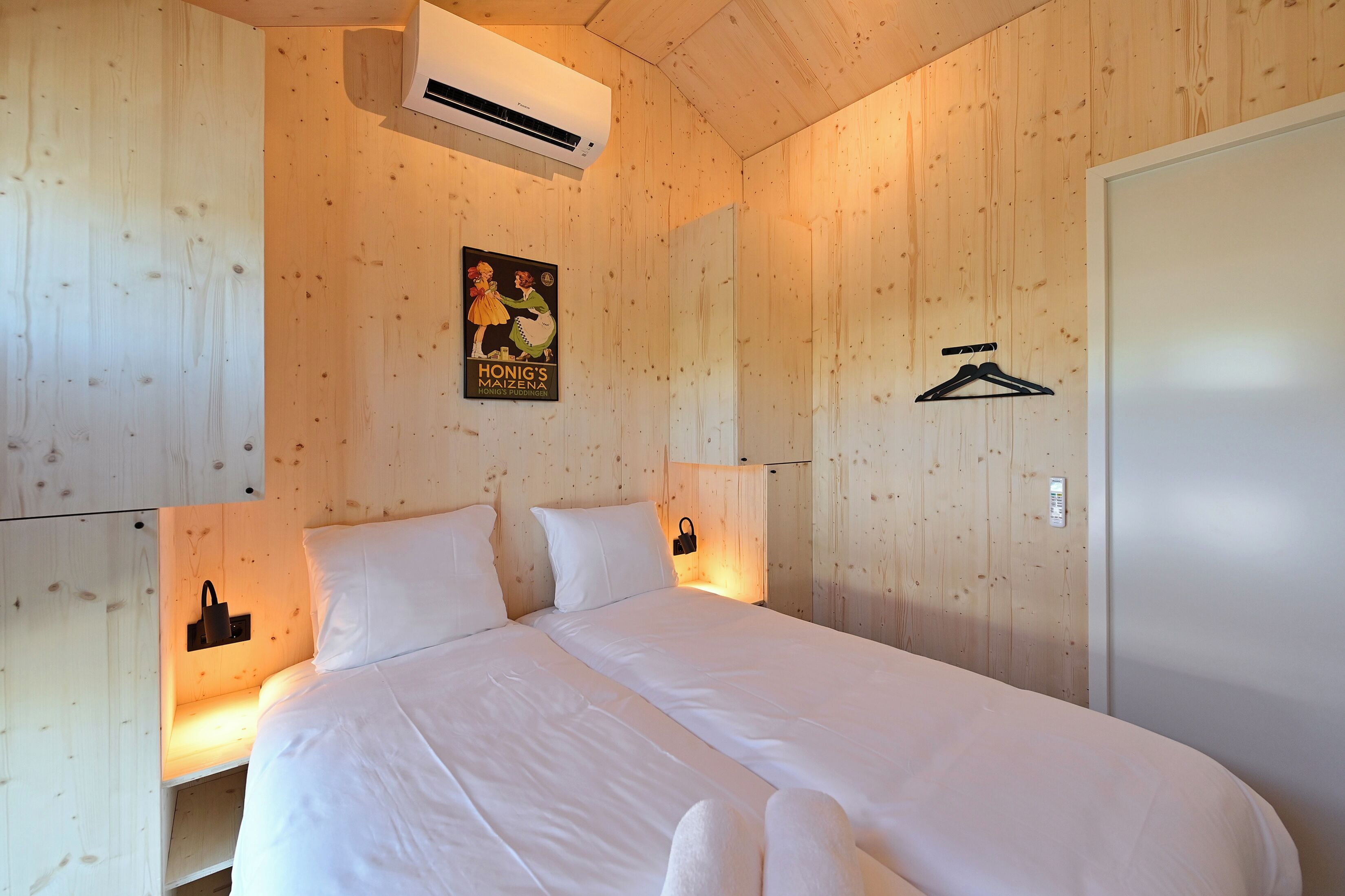 A bedroom with a wooden wall, a bed with white bedding, an air conditioning unit, a poster, a coat hanger, and a thermostat.