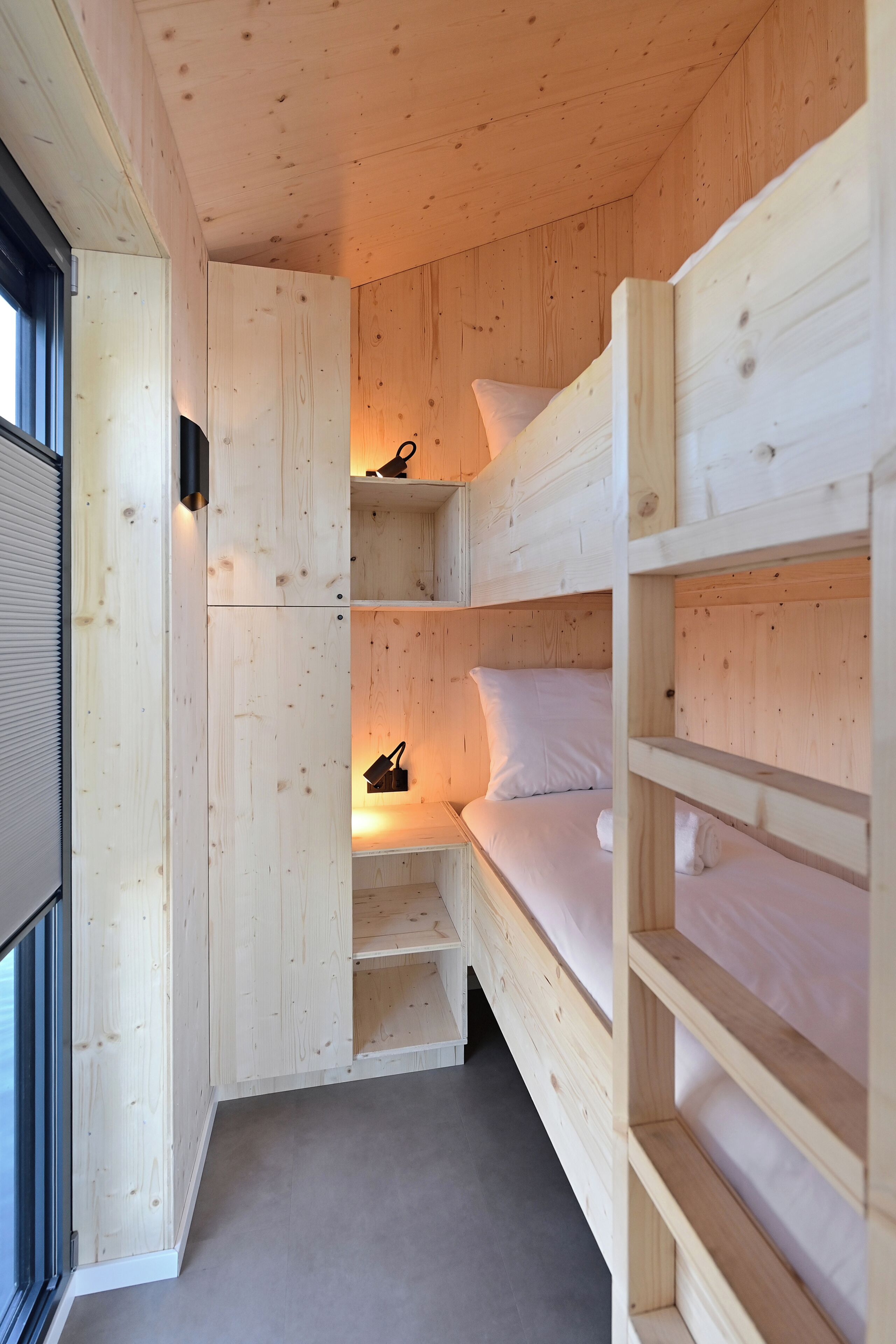 Habitación de madera pequeña con literas, estanterías integradas y una ventana.