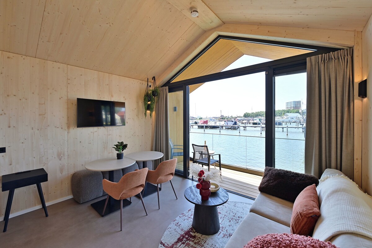 Un interior moderno con techo de madera, un televisor de pantalla plana, una zona de comedor con sillas y mesa, un sofá y vistas a un puerto deportivo.