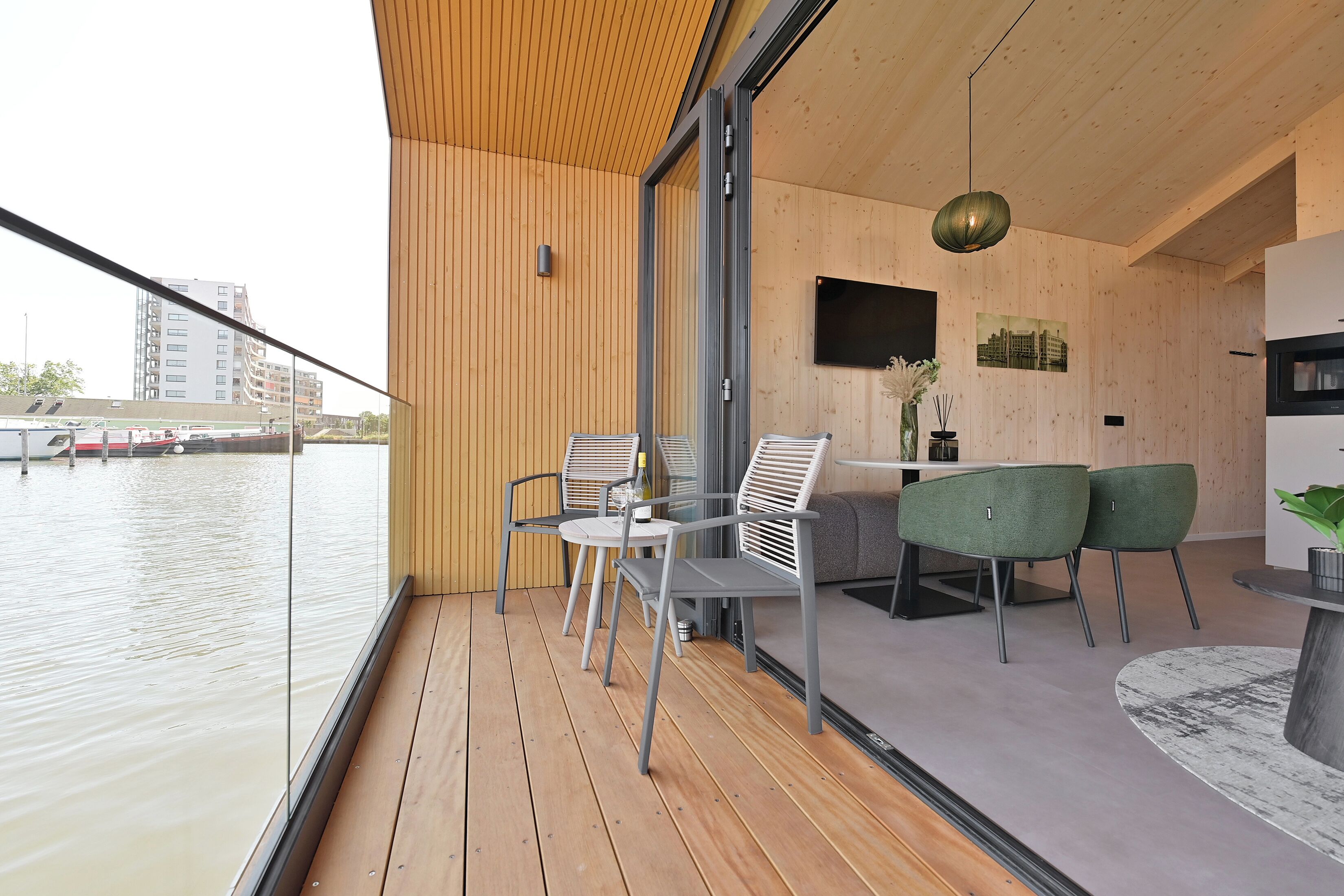 Un interior moderno con terraza de madera, zona de comedor con sillas y mesa, y cocina con televisor y plantas.