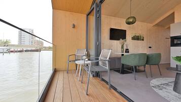Un interior moderno con terraza de madera, zona de comedor con sillas y mesa, y cocina con televisor y plantas.