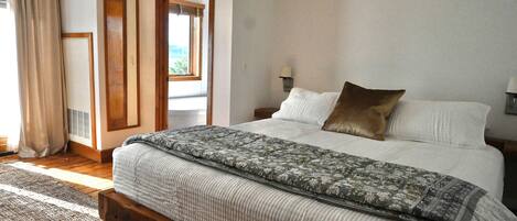 Une chambre à coucher avec un lit en bois, une couvre-lit à motifs, une vue sur l’extérieur par une porte ouverte et une fenêtre avec des rideaux.