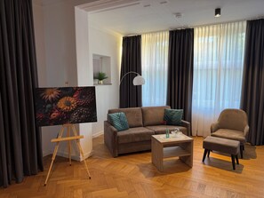 A living room with a sofa, armchair, coffee table, and an easel with a painting.