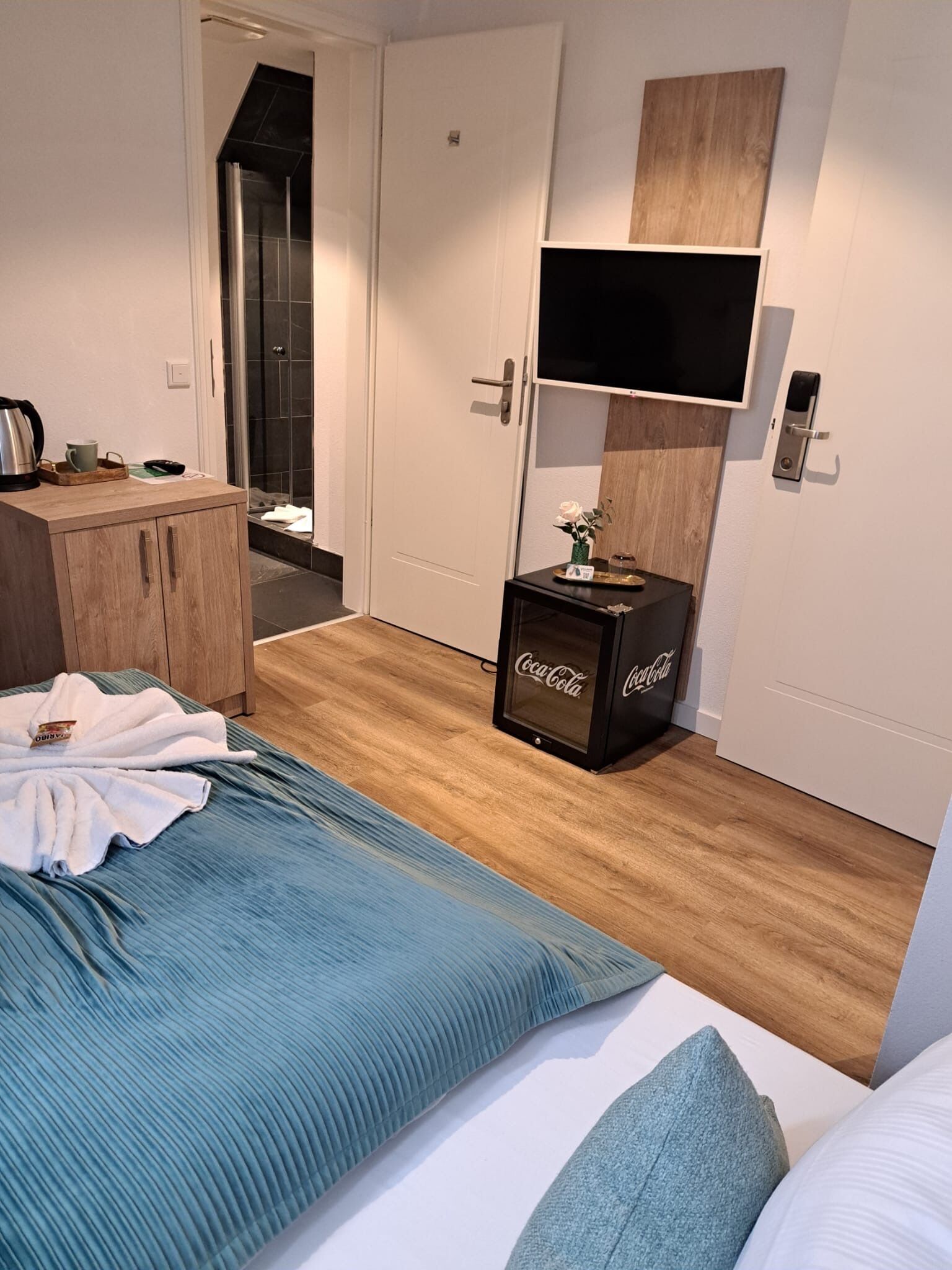 A hotel room with a bed, a wooden dresser, a television mounted on the wall, and a small table with a Coca-Cola branded item.