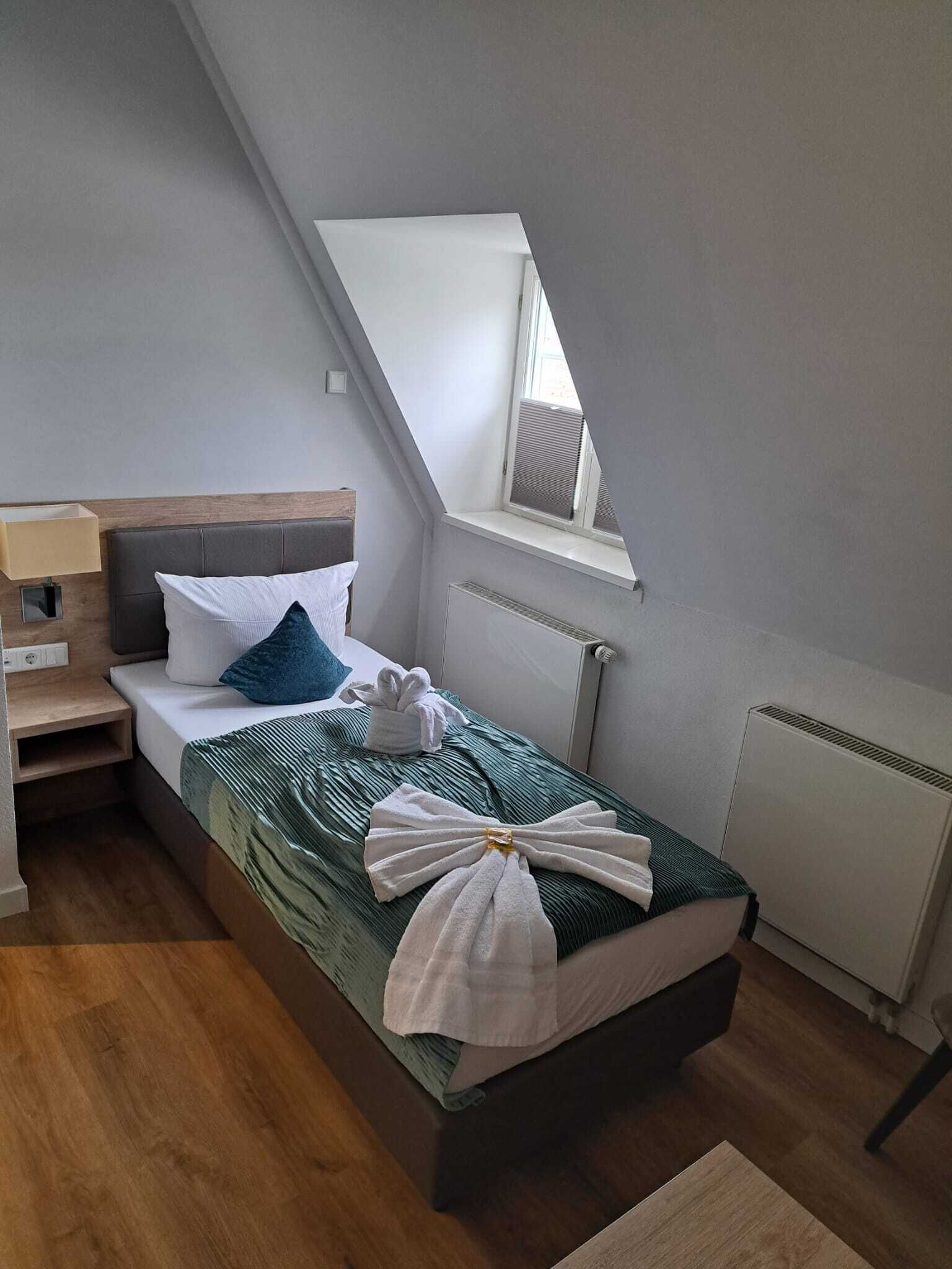 A single bed with a green bedspread, white pillows, and a towel arrangement. A small bedside table with a lamp is visible. The room has a sloped ceiling with a window and a radiator.