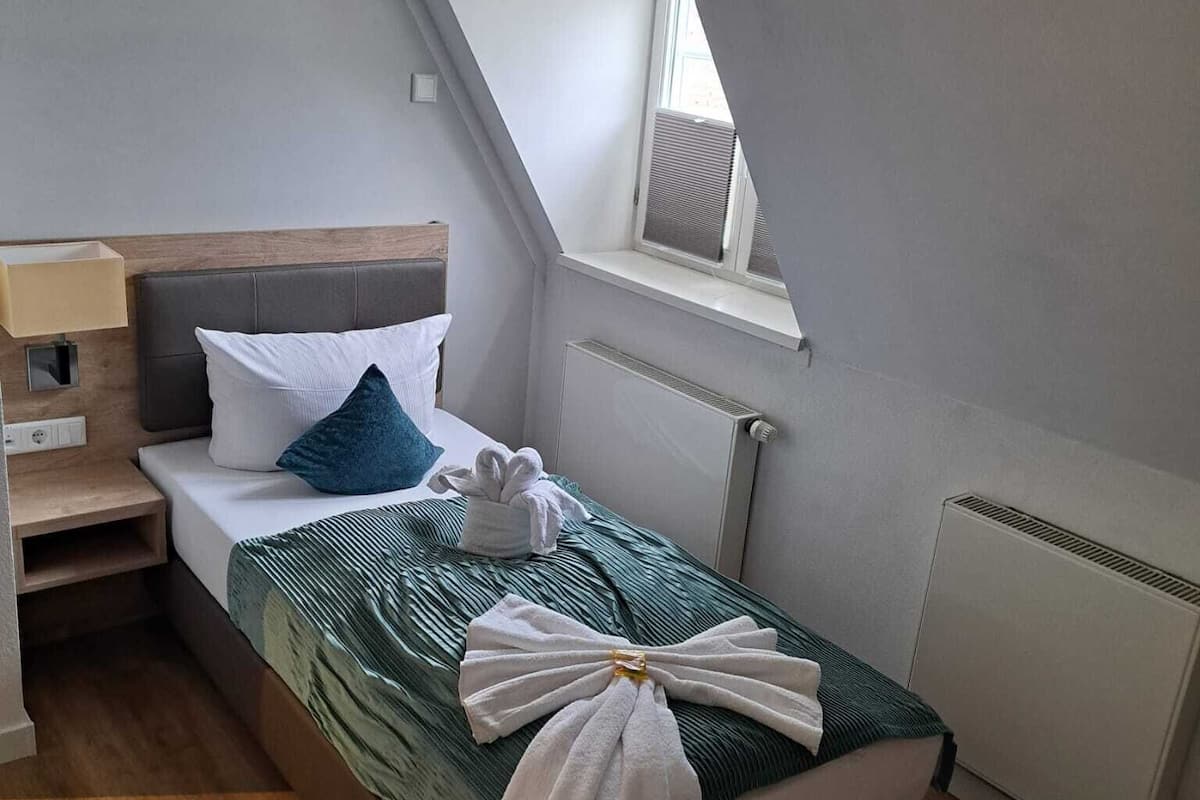 A single bed with a green bedspread, white pillows, and a towel arrangement. A small bedside table with a lamp is visible. The room has a sloped ceiling with a window and a radiator.