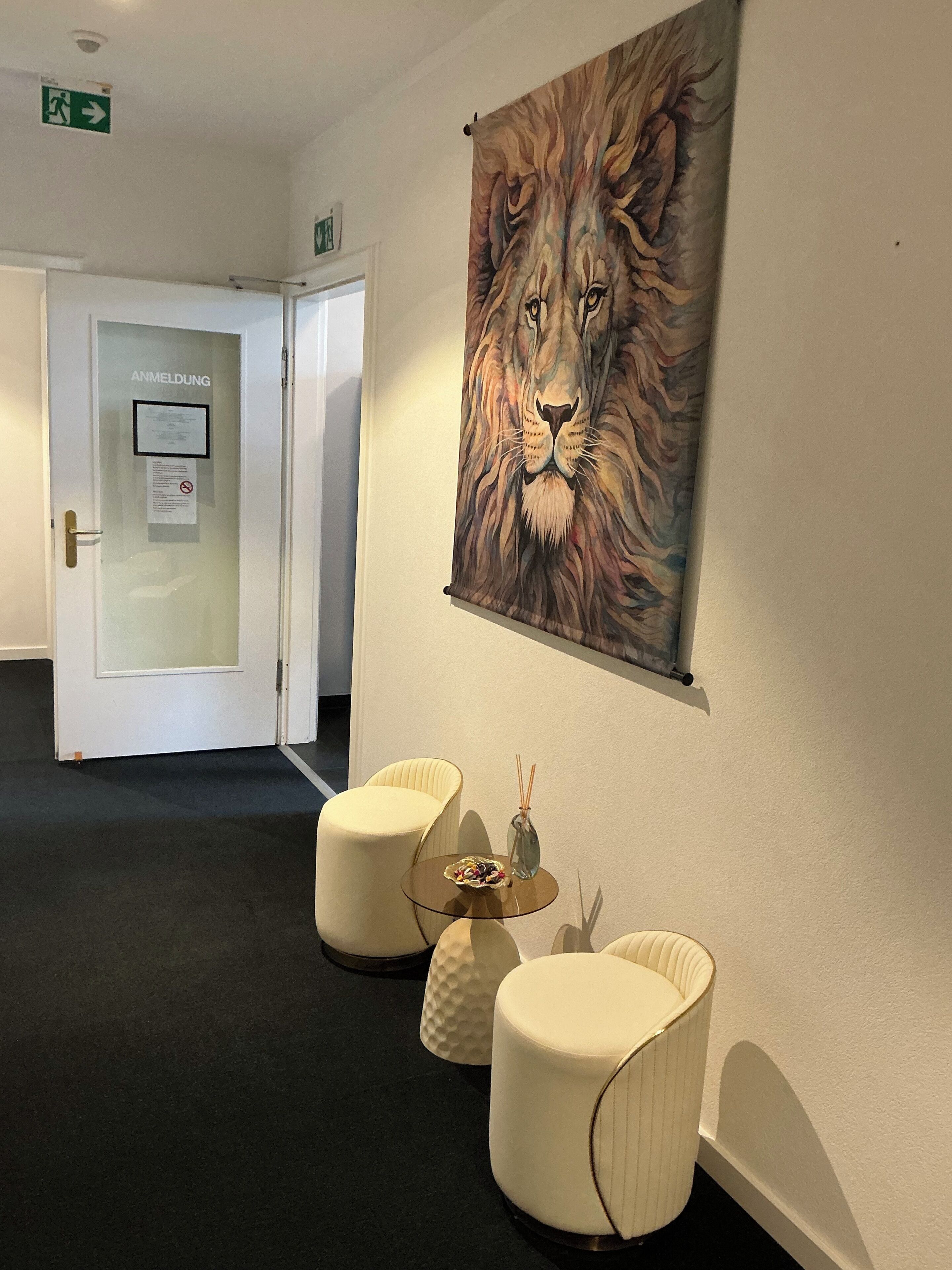 Eine Hotelhalle mit Glastür, auf der 'ANMELDUNG' steht, einem gerahmten Löwenbild und zwei weißen Sesseln mit einem kleinen Tisch dazwischen.