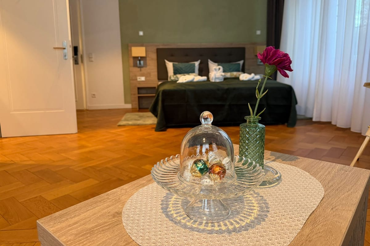 Una habitación de hotel con una cama, una mesita de noche con una cúpula de vidrio y objetos decorativos, un jarrón verde con una flor y un suelo de madera.