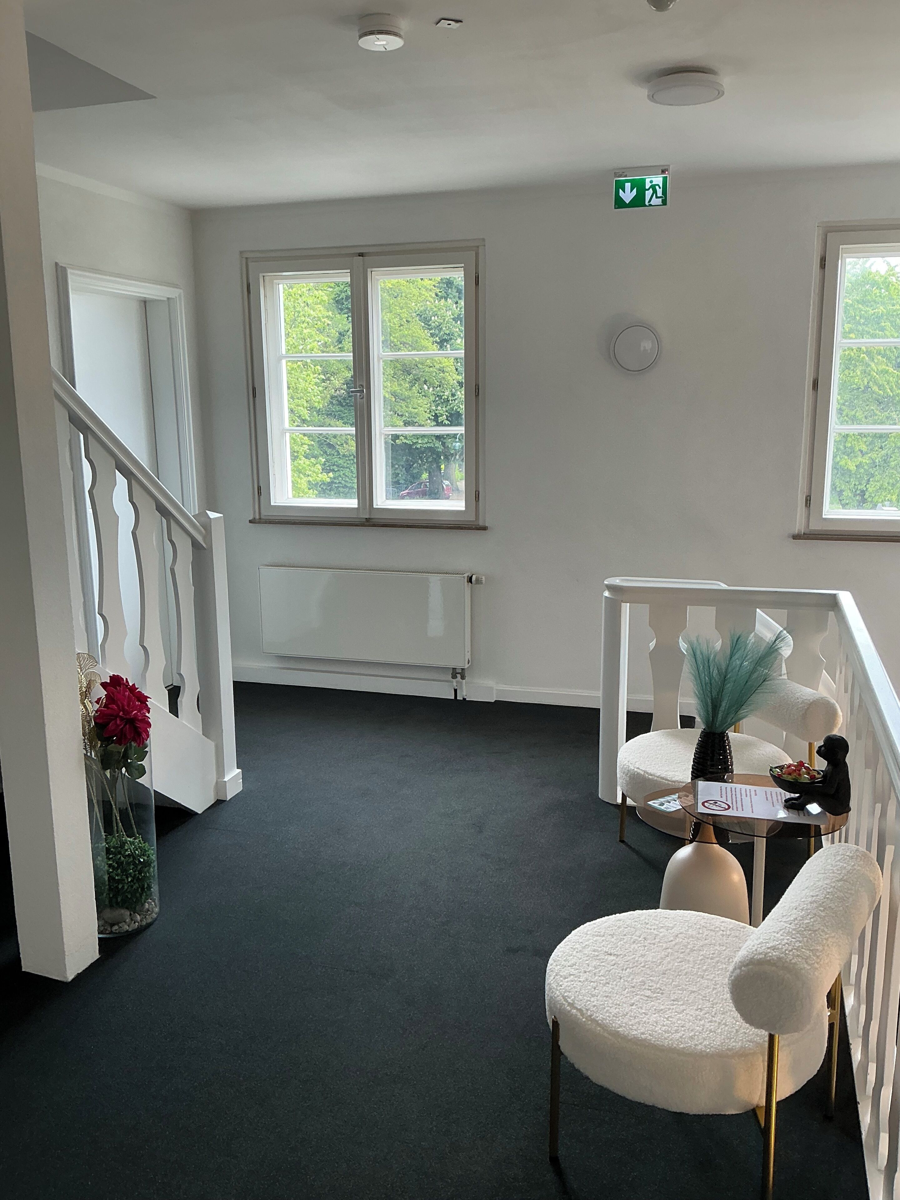A room with a radiator, a white chair, a small table with a vase, and a window with a view of greenery.