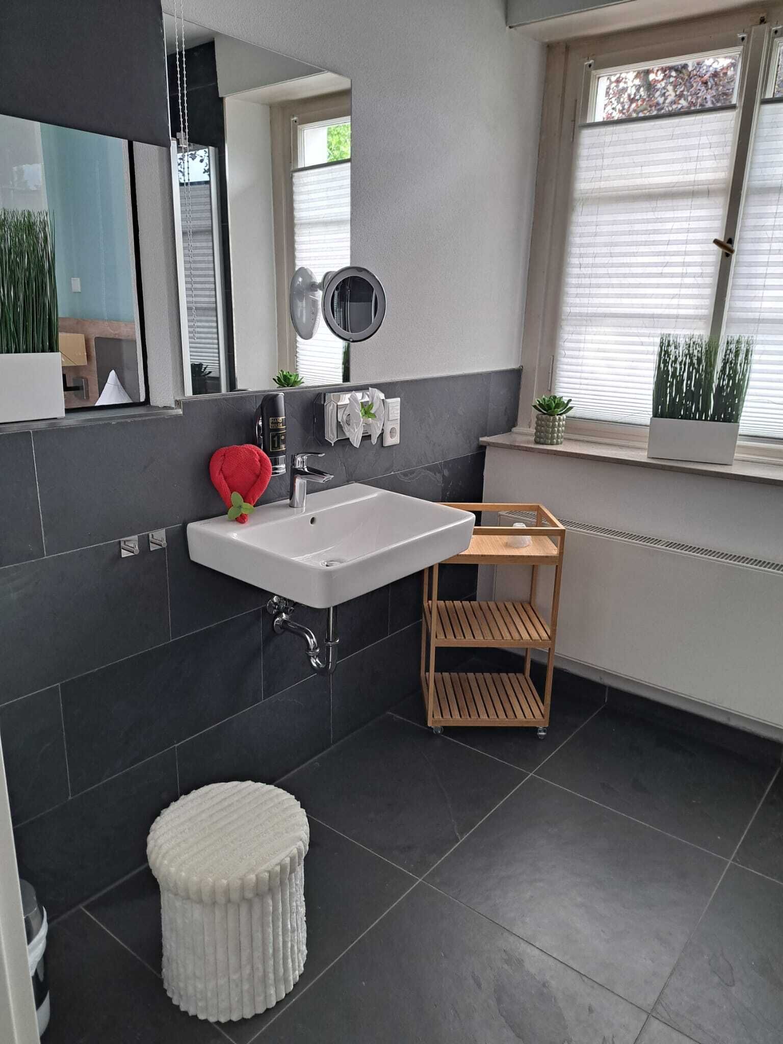 A modern bathroom with a white sink, a round mirror, a small stool, and a wooden shelf.