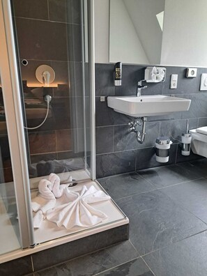 A modern bathroom with a glass shower enclosure, a white sink, and dark tiled walls.