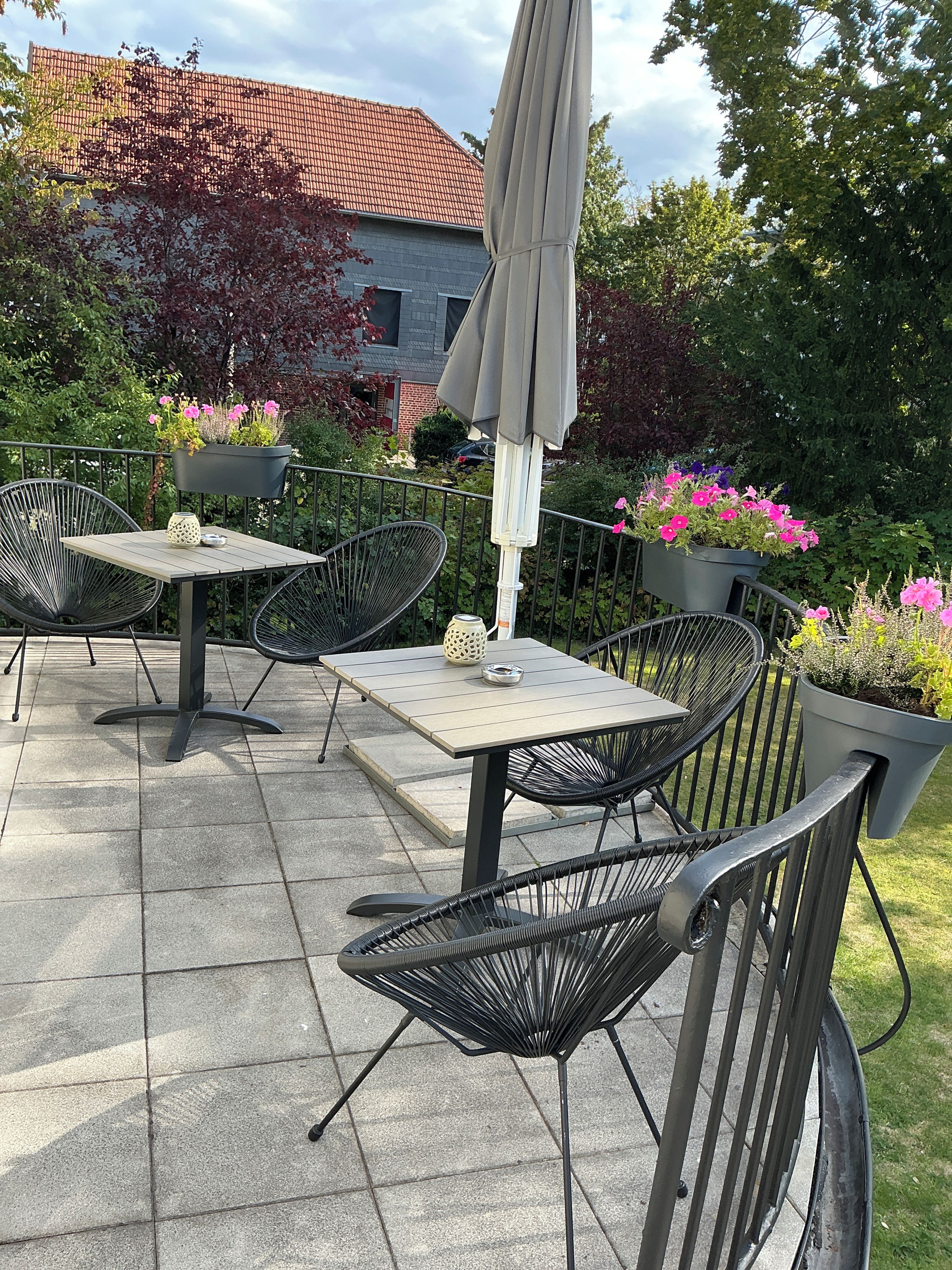 Eine Terrasse im Freien mit Tisch und Stühlen, umgeben von Blumentöpfen und einem Gebäude im Hintergrund.