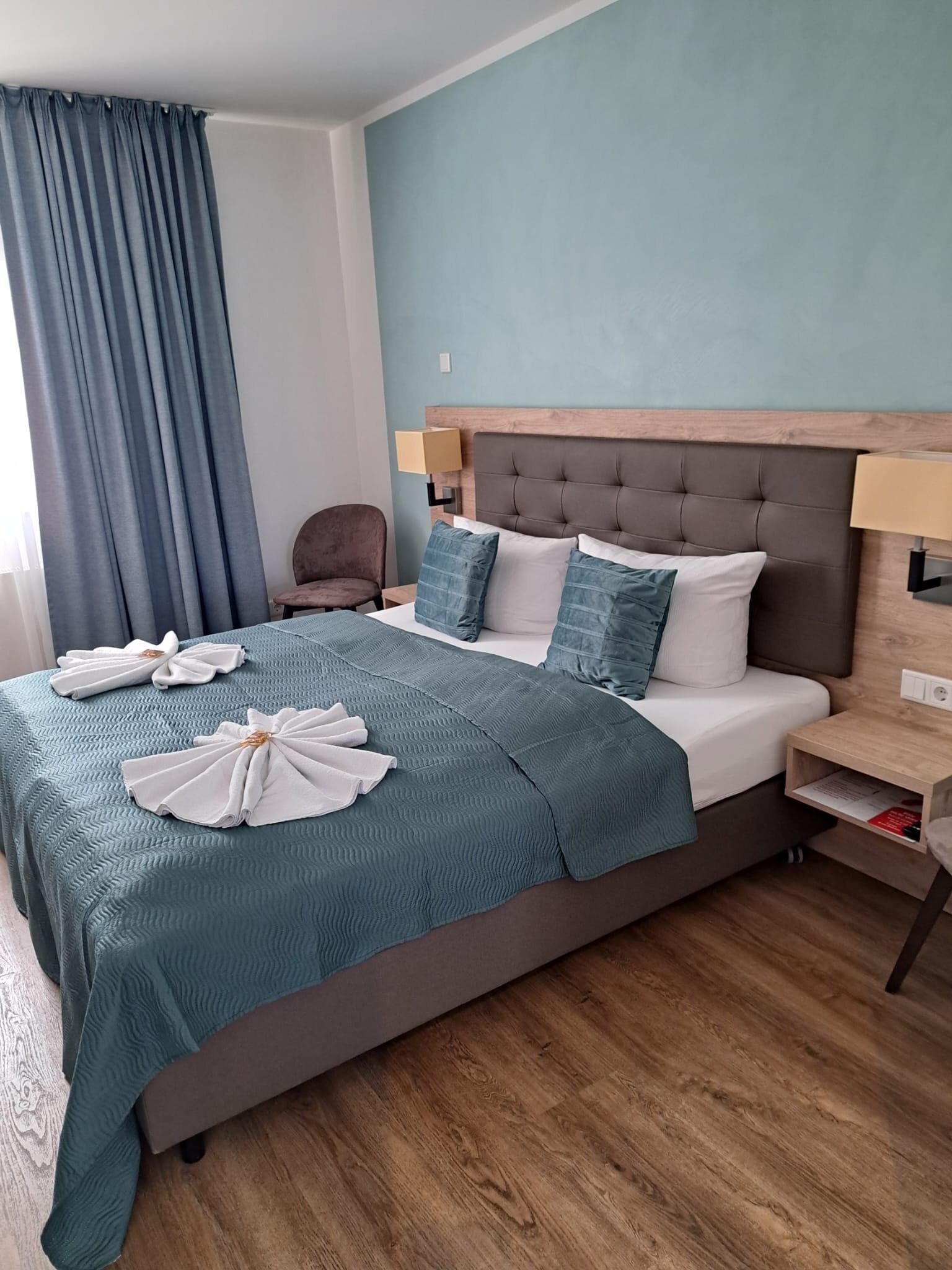 A neatly made bed with a teal bedspread and white pillows, a wooden headboard, a bedside table with a lamp, and a chair.