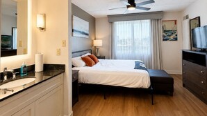Ein Hotelzimmer mit Bett, Schreibtisch, Fernseher, Deckenventilator und Badezimmer mit Waschbecken und Spiegel.