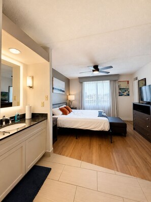 A hotel room with a bed, a desk, a TV, a ceiling fan, and a bathroom with a sink and mirror. -  Elite Dallas Medical District Getaway with Pool View (Dallas)