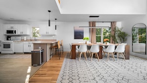A modern kitchen with white cabinets, a wooden dining table, and a view of greenery outside. - Platte River Sauna, Hot Tub & Kayaks, Near Dunes (Honor)