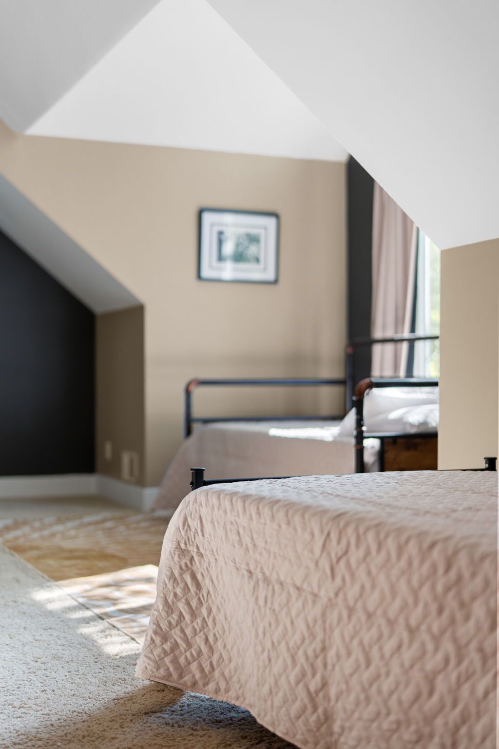 A bedroom with a sloped ceiling, a bed with a quilted cover, a framed picture on the wall, and a window with curtains.