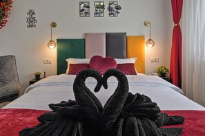 Habitación de hotel con una cama adornada con una presentación de toallas en forma de corazón, cabecera verde y gris, y dos lámparas fijadas a la pared.