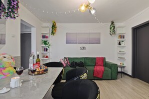 Una sala de estar moderna con un sofá verde, una mesa de comedor con sillas, una mesita auxiliar pequeña y una pared con un cuadro enmarcado.