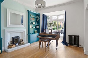 Een kamer met een piano, een open haard, een boekenkast en uitzicht door een groot raam.