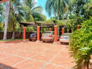 A covered parking area with three cars parked under a structure supported by red pillars, surrounded by lush greenery and palm trees.