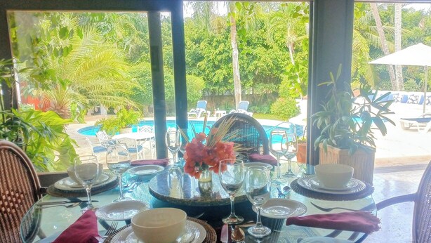 A dining table with glassware and a bowl, surrounded by wicker chairs, with a view of a pool and greenery outside. - Coral Suite at Casa de Campo Resort & Villas Golf Playa (La Romana)