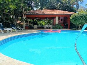 En swimmingpool med fliset kant, liggestole, et overdækket opholdsområde og en palme.