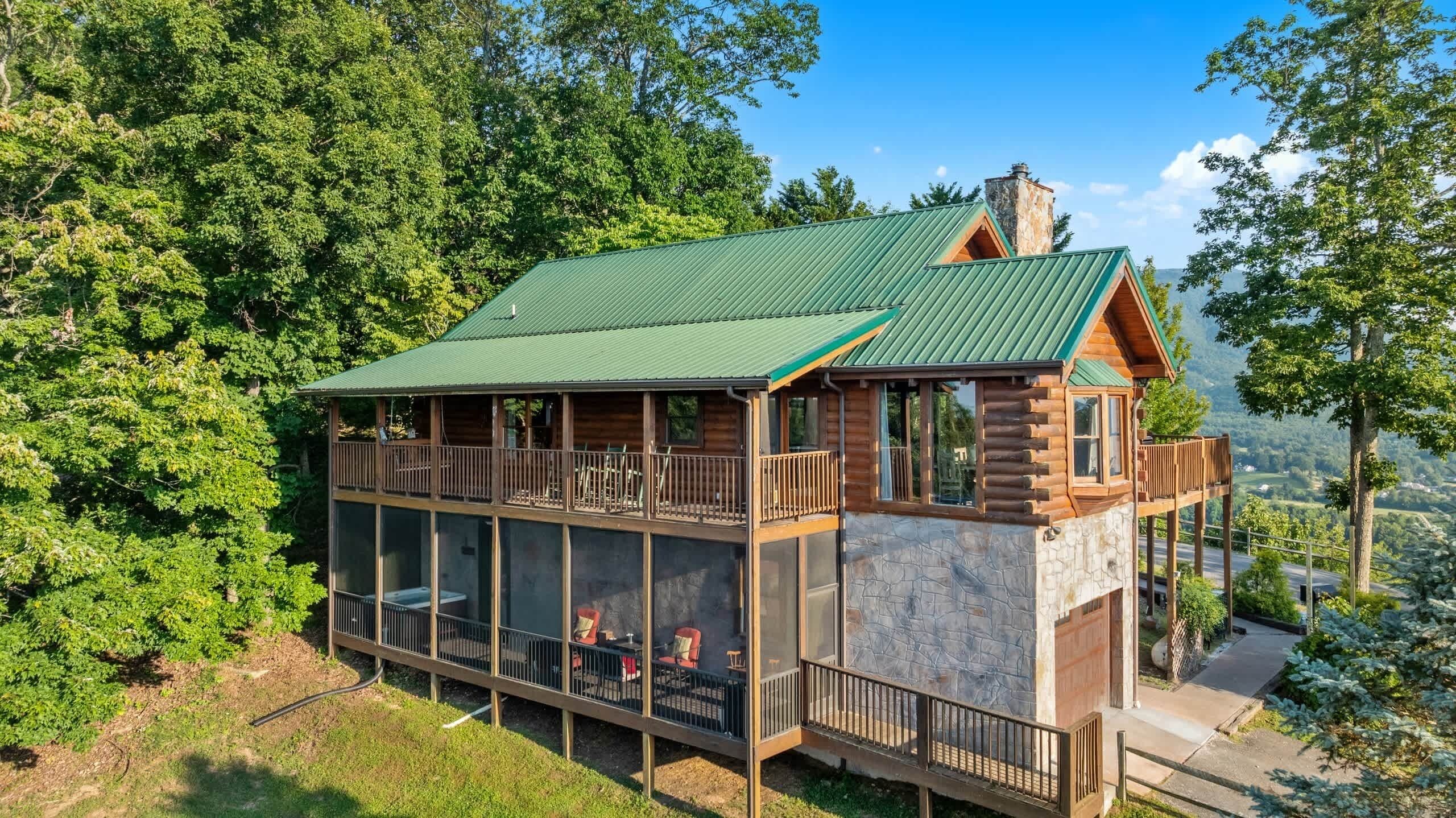 A two-story log cabin with a green metal roof, large windows, and a stone facade.