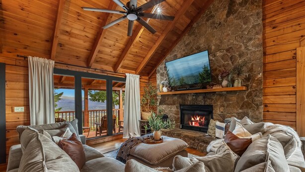 A cozy living room with a stone fireplace, a flat-screen TV mounted on the wall, and a comfortable seating area with a sofa and chairs. - *New* Golf Sim Cabin | Views | Hot Tub | Fire Pit (Sevierville)