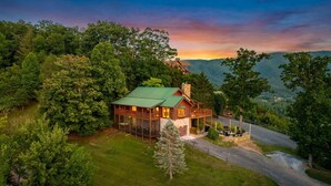 A log cabin with a green roof, surrounded by dense forest, and a clear view of the mountains in the background. - *New* Golf Sim Cabin | Views | Hot Tub | Fire Pit (Sevierville)