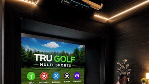 A dark room with a large screen displaying 'TRU GOLF MULTI SPORTS' and icons for various sports. There's a golf bag and a small table with a plant. - *New* Golf Sim Cabin | Views | Hot Tub | Fire Pit (Sevierville)