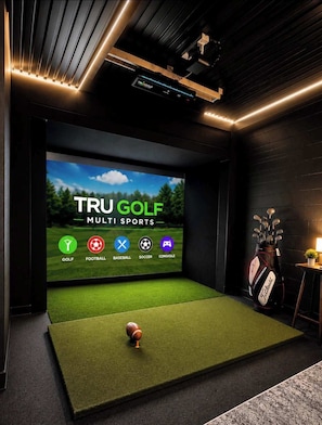 A dark room with a large screen displaying 'TRU GOLF MULTI SPORTS' and icons for various sports. There's a golf bag and a small table with a plant. - *New* Golf Sim Cabin | Views | Hot Tub | Fire Pit (Sevierville)