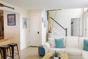 Ein Wohnzimmer mit einem weißen Sofa, einem Couchtisch, einem blauen Hocker, einem hölzernen Barhocker und einer Treppe mit blauer Geländer.