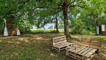 Un espace extérieur aménagé dans un style rustique, avec des bancs en bois et une petite table, entouré d’arbres et d’un champ d’herbe.
