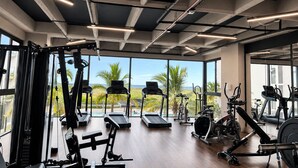 Ein Fitnessstudio mit Trainingsgeräten, großen Fenstern und Blick auf Palmen.