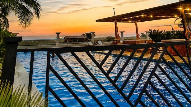 Blick über den Pool bei Sonnenuntergang, mit Palmen und einem überdachten Bereich mit Beleuchtung.