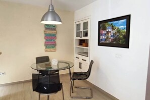 Ein Essbereich mit einem Glastisch, zwei schwarzen Stühlen, einer Pendelleuchte, einem weißen Schrank und einem gerahmten Bild an der Wand.