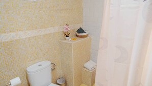 Un baño con inodoro, un cubo de basura pequeño, lavamanos y una planta decorativa en un estante.