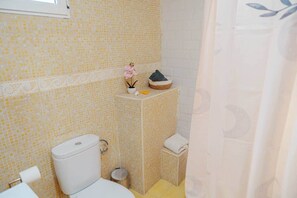 Ein Badezimmer mit Toilette, einem kleinen Mülleimer, einem Waschbecken und einer Zimmerpflanze auf einem Regal.