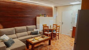 Una sala de estar con sofá, mesa de centro y sillas, un muro decorativo de madera, y una zona de cocina al fondo.