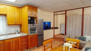 Cocina compacta con armarios de madera, microondas, horno y refrigerador. Hay un comedor con mesa y sillas, y una sala de estar con sofá y televisor.