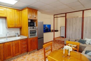 Eine kompakte Küche mit Holzausstattung, Mikrowelle, Backofen und Kühlschrank. Es gibt einen Essbereich mit Tisch und Stühlen sowie ein Wohnzimmer mit Sofa und Fernseher.