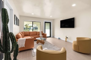 Ein Wohnzimmer mit einer Couch, Sesseln, einem Couchtisch, einem Kaktus und einem an der Wand befestigten Fernseher.