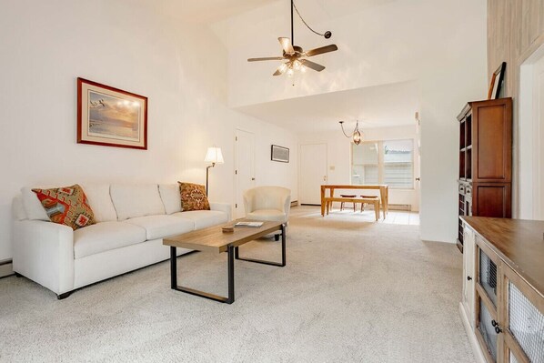 A living room with a white sofa, a wooden coffee table, a white armchair, a dining table with chairs, a ceiling fan, and a kitchen area with wooden cabinets.