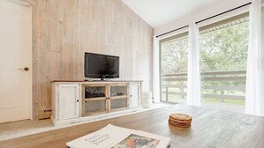 A room with a wooden floor, a television on a cabinet, a large window with a view of trees, and a magazine on the table.