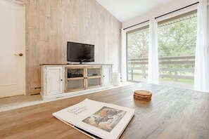 A room with a wooden floor, a television on a cabinet, a large window with a view of trees, and a magazine on the table. - Cozy Condo Ranch at Roaring Fork (Carbondale)
