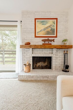 A white brick fireplace with a wooden mantel, a framed painting above it, and a view of greenery outside. - Cozy Condo Ranch at Roaring Fork (Carbondale)