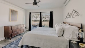 Un dormitorio con una cama, un ventilador de techo, vistas por la ventana, un aparador de madera y una alfombra estampada.