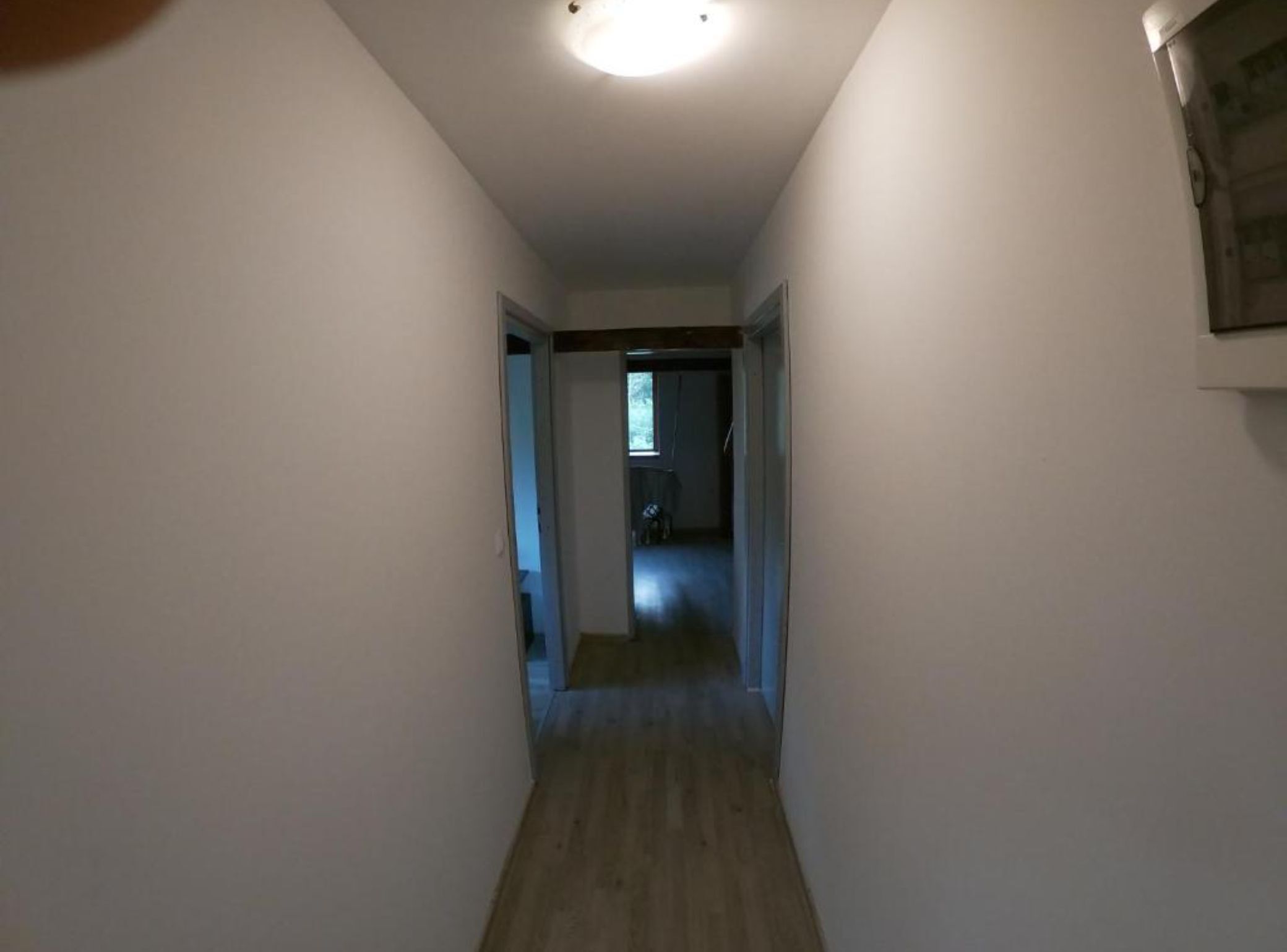 A narrow hallway with white walls and wooden flooring, leading to a room with a door.